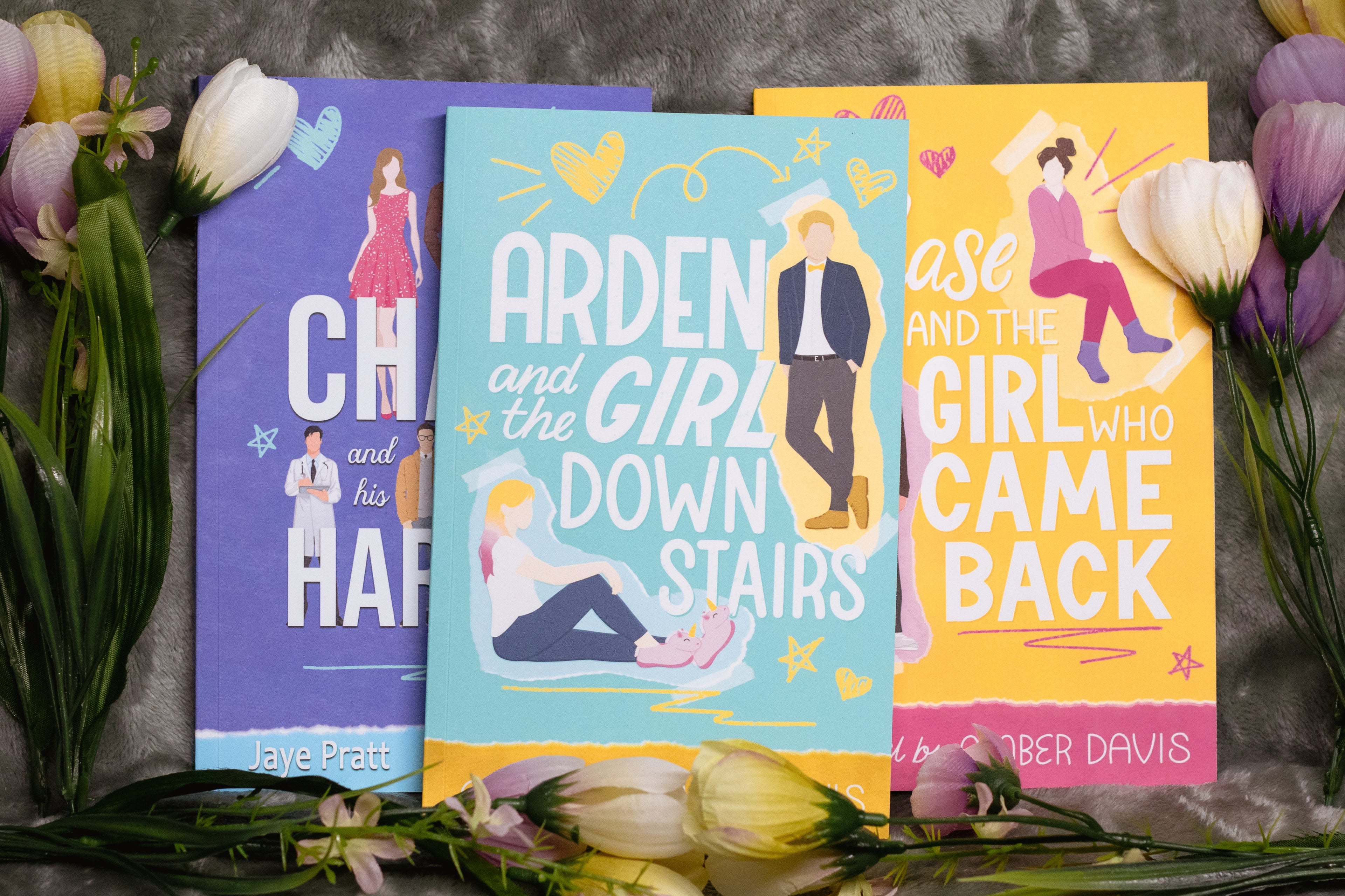 "Three romance books displayed on a soft grey fabric background, surrounded by purple, white, and yellow tulips. The books are titled Chad and His Harem by Amber Davis and Jaye Pratt, Arden and the Girl Downstairs, and Chase and the Girl Who Came Back by Amber Davis. Each cover features a colourful illustrated design with silhouettes of the main characters in various poses, along with playful doodles and typography."